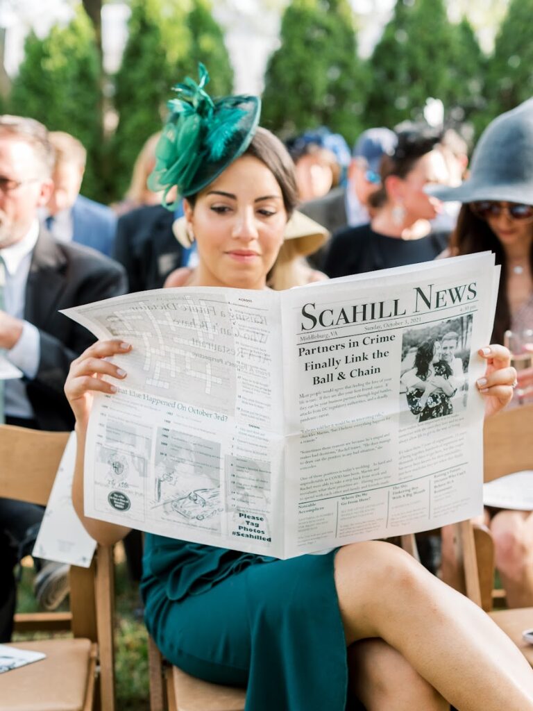 a woman in a green dress and fascinator reads the bride and grooms custom newspaper while waiting for the ceremony to begin