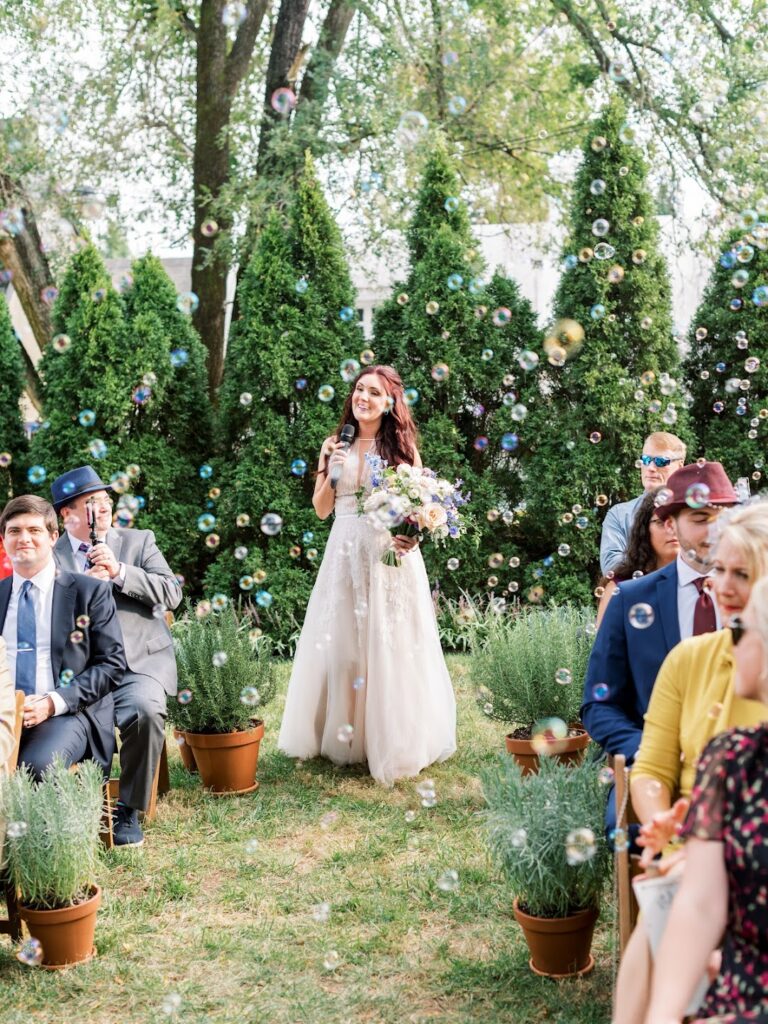 a bride walks down an aisle flanked with large potted herbs, singing into a microphone while bubbles float around her.