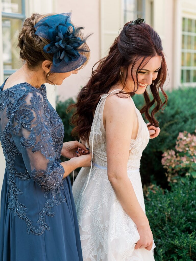 A bride being zipped into her wedding dress by her mom dressed in a formal blue dress and matching blue fascinator