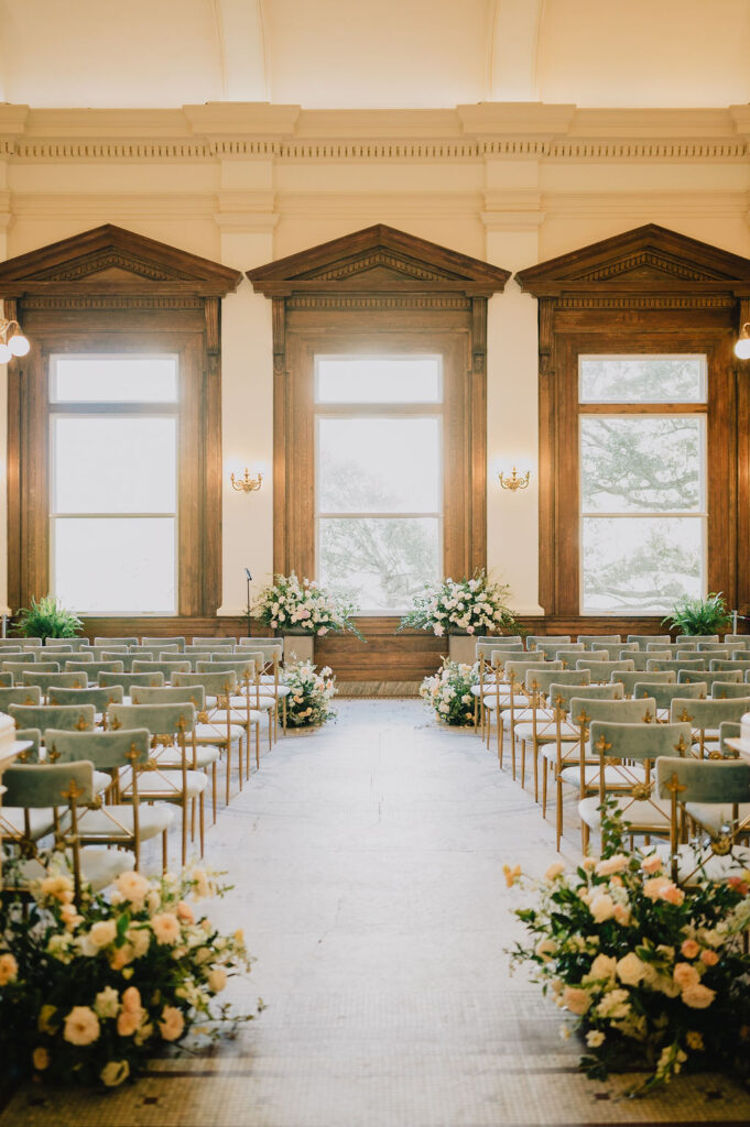 A symmetrical wide shot of a luxury wedding ceremony setup in the Gibbes Museum Rotunda featuring sage green velvet chairs and lush floor floral arrangements in ivory and peach.