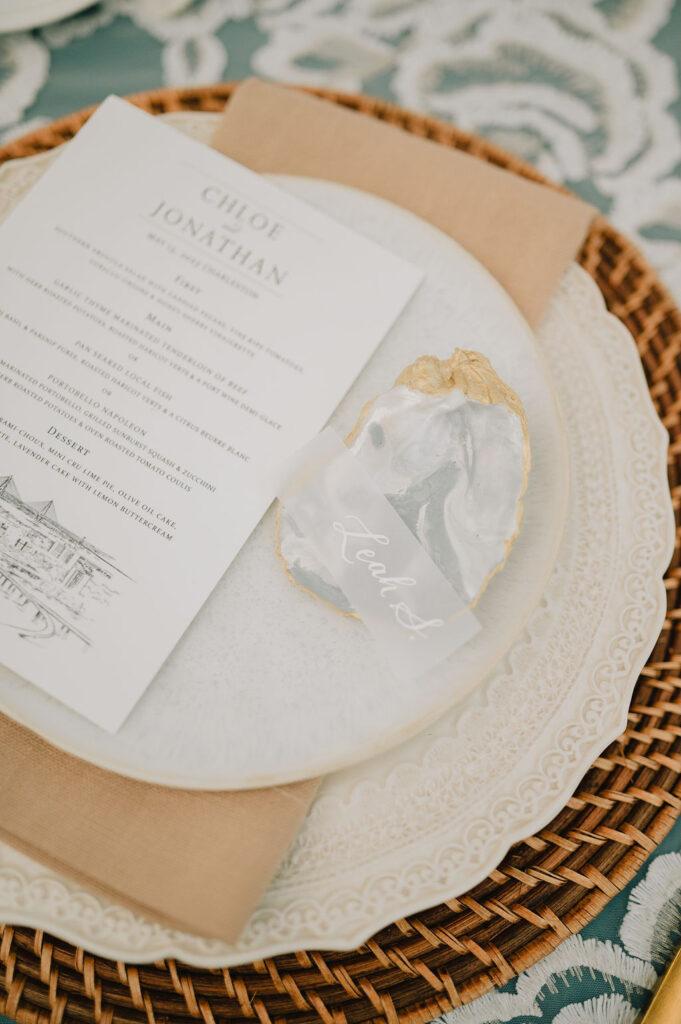 Modern Lowcountry wedding table decor featuring hand-painted gold oyster shell place cards, rattan chargers, and stone-colored plates at a Charleston museum reception.