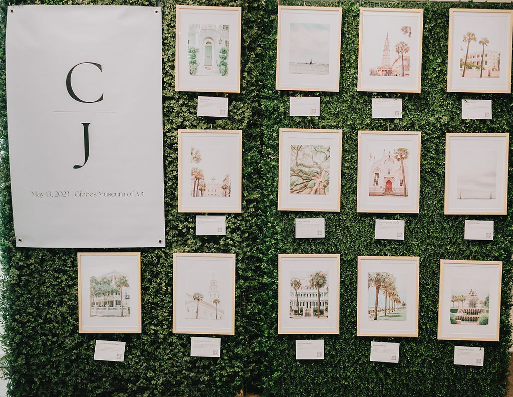 A creative wedding escort card display featuring a gallery wall of local Charleston art prints with QR codes for an interactive guest experience at a museum wedding.